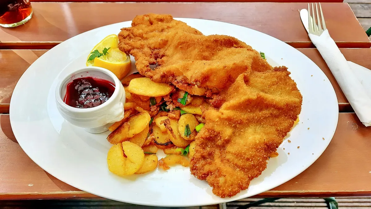 Original phone photo: a schnitzel plate with fried potatoes, a lemon wedge and a pot of berry sauce on an outdoor timber table, with a glass and cutlery at the edges